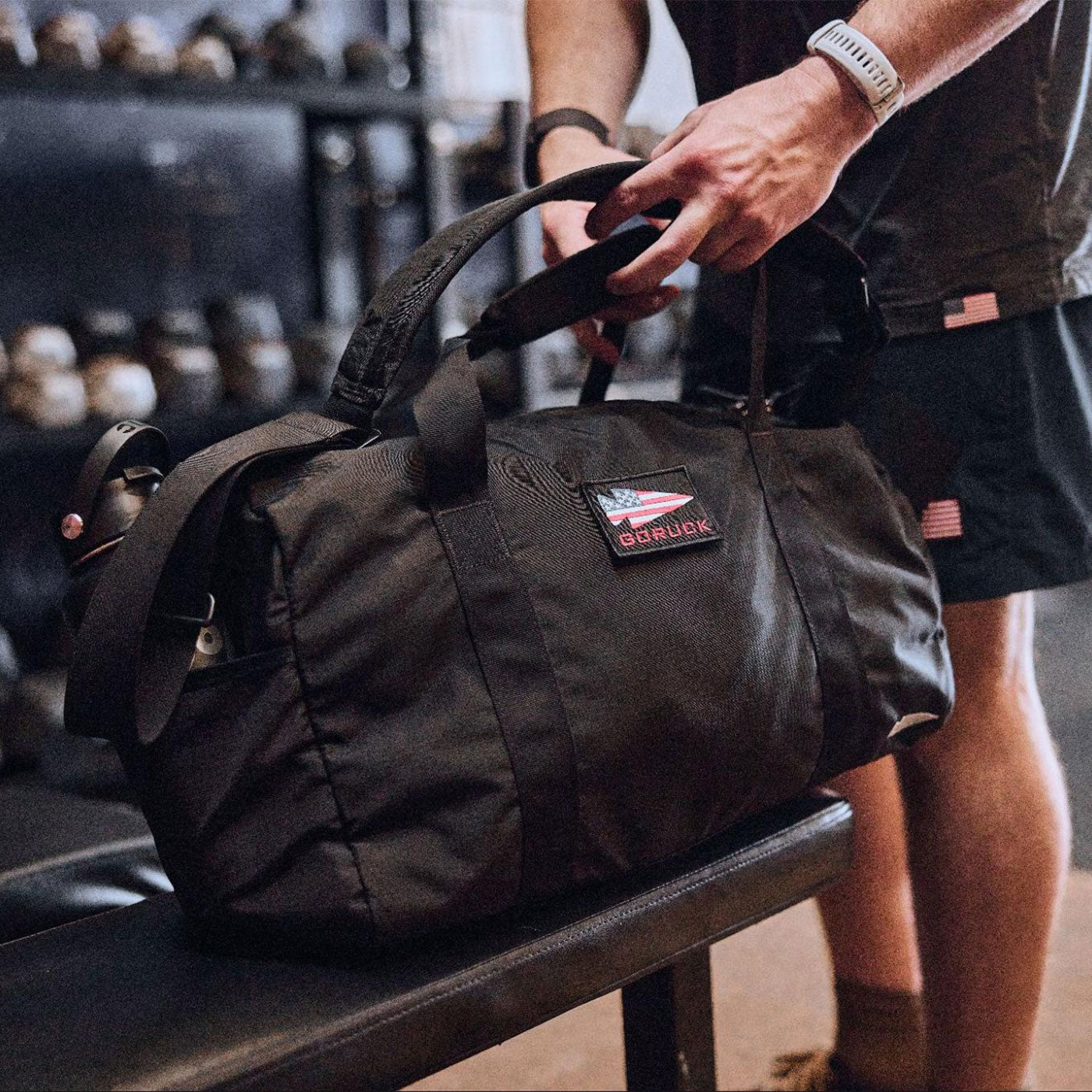 Person placing a black gym bag with a GORUCK patch on a bench in a gym, weights visible in the background.