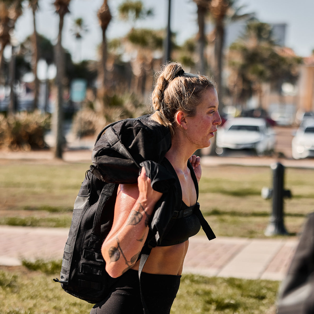 Woman exercising outdoors, carrying a heavy black sandbag on her shoulders, with palm trees in the background.