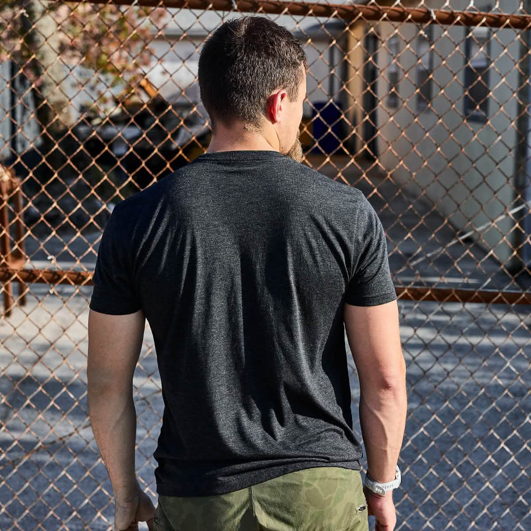 A man wears the Subtle Spearhead Tee - Tri-Blend by vendor-unknown, facing a chain-link fence outdoors on a sunny day, with its subtle spearhead emblem nodding to his Special Forces spirit.
