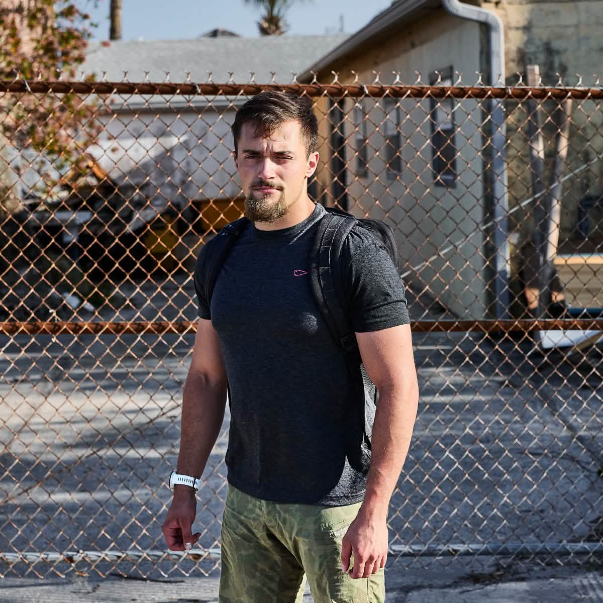 A man wearing the Subtle Spearhead Tee - Tri-Blend by vendor-unknown stands before a chain-link fence on a sunny day, channeling the spirit of Special Forces.