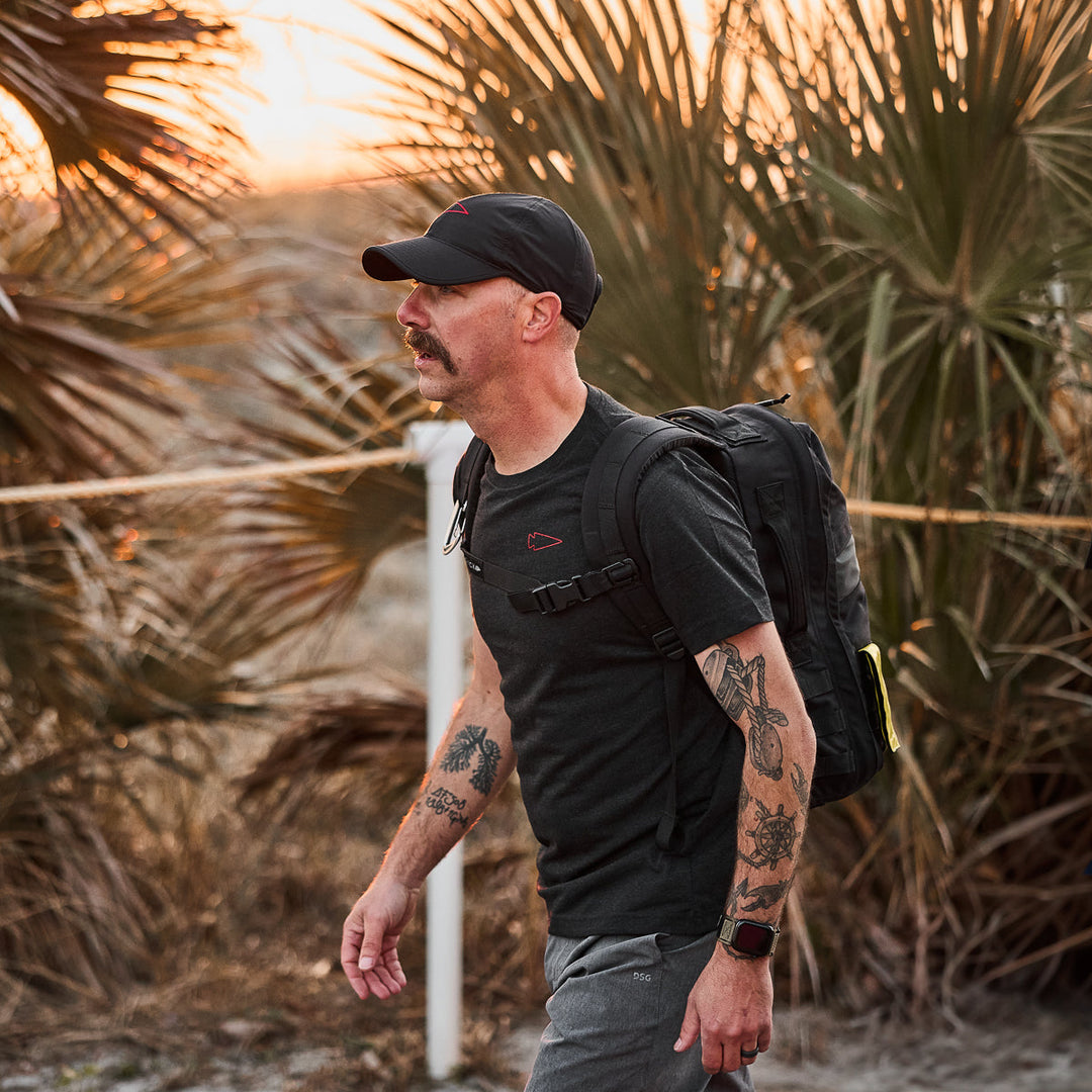 Tattooed man with mustache walks outdoors at sunset, wearing a black cap, backpack, and the vendor-unknown Subtle Spearhead Tee - Tri-Blend.
