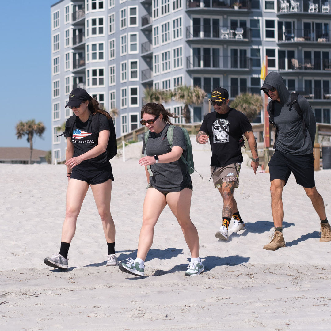 Four people in Subtle Spearhead Tee - Tri-Blend shirts from vendor-unknown walk along a sandy beach, tall modern buildings behind them—capturing the GORUCK spearhead spirit of Special Forces camaraderie.