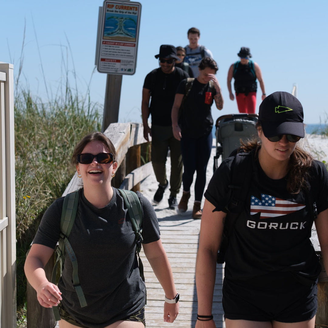 A group wearing vendor-unknown Subtle Spearhead Tee - Tri-Blend shirts with backpacks walk along a wooden path toward the beach on a sunny day.