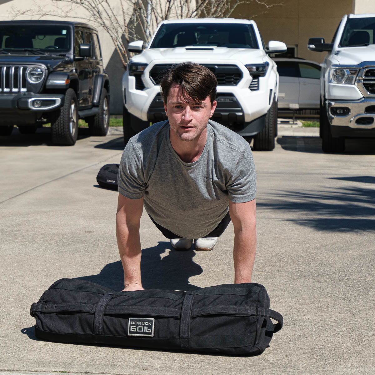 Man in heather grey GORUCK tee doing push-ups outdoors with GORUCK sandbag, vehicles behind
