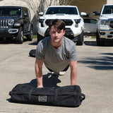 Man in heather grey GORUCK tee doing push-ups outdoors with GORUCK sandbag, vehicles behind