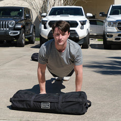 Man in heather grey GORUCK tee doing push-ups outdoors with GORUCK sandbag, vehicles behind