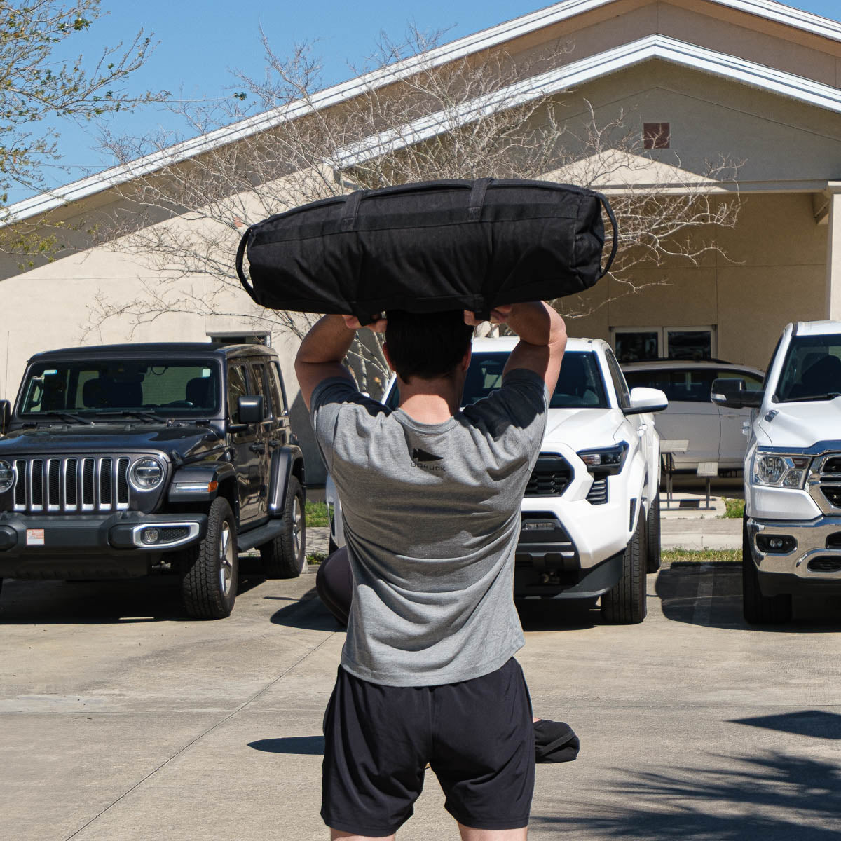 Man in GORUCK gray tee rucking outdoors, lifting sandbag overhead in parking lot