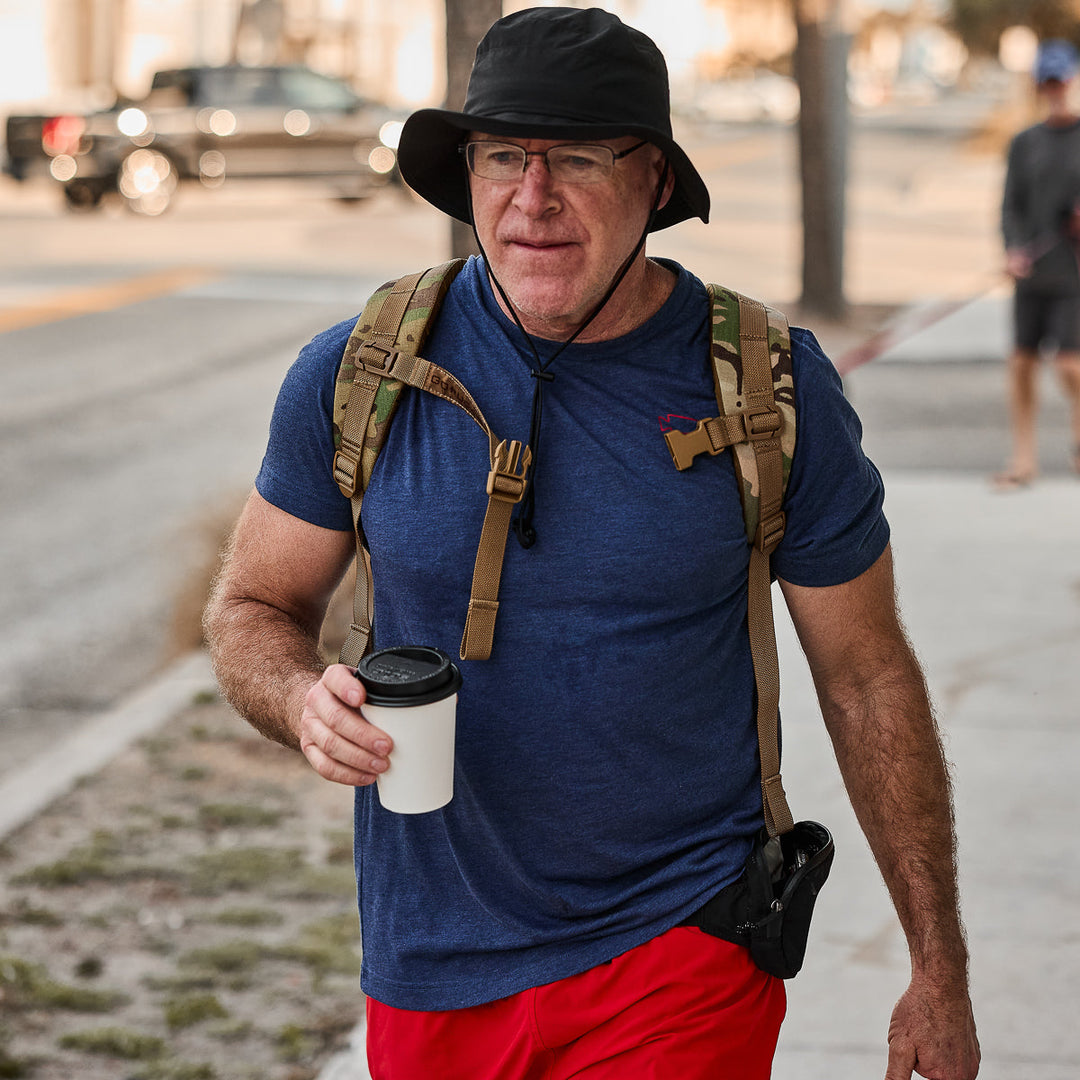 A man walks outside wearing the Subtle Spearhead Tee - Tri-Blend by vendor-unknown, paired with red shorts, a black hat, a backpack featuring a GORUCK spearhead patch, and holding a coffee cup.