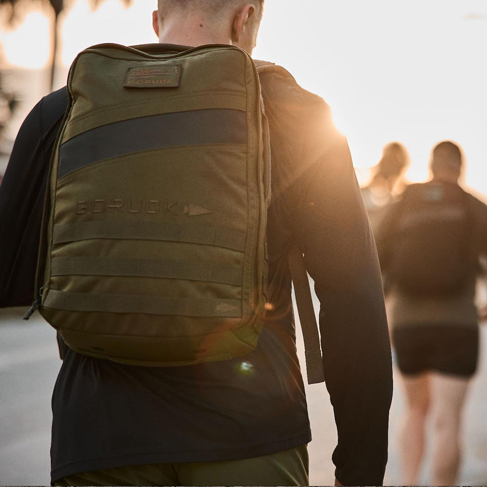 Person wearing an olive backpack, walking outdoors at sunrise, followed by two others with backpacks.