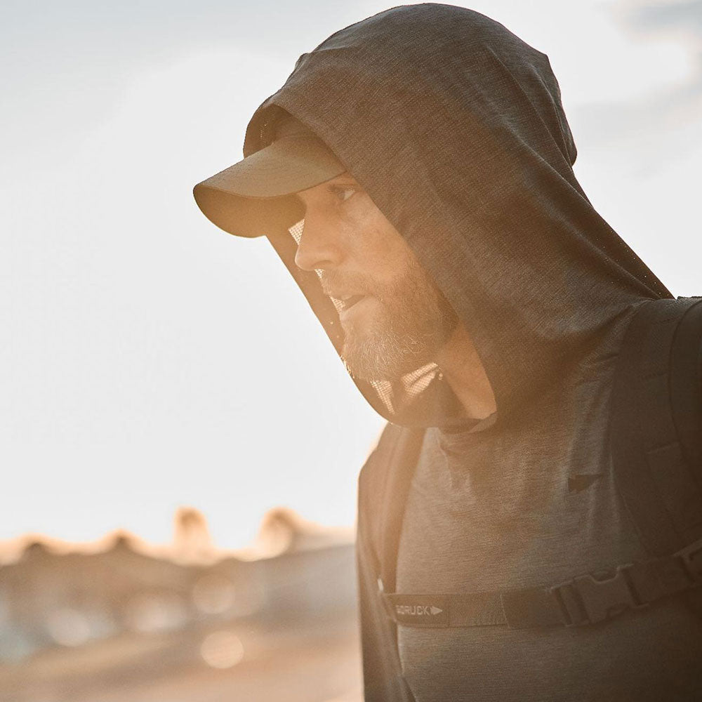 Man in a hooded shirt and cap, outdoors, wearing a backpack, with sunlight in the background.