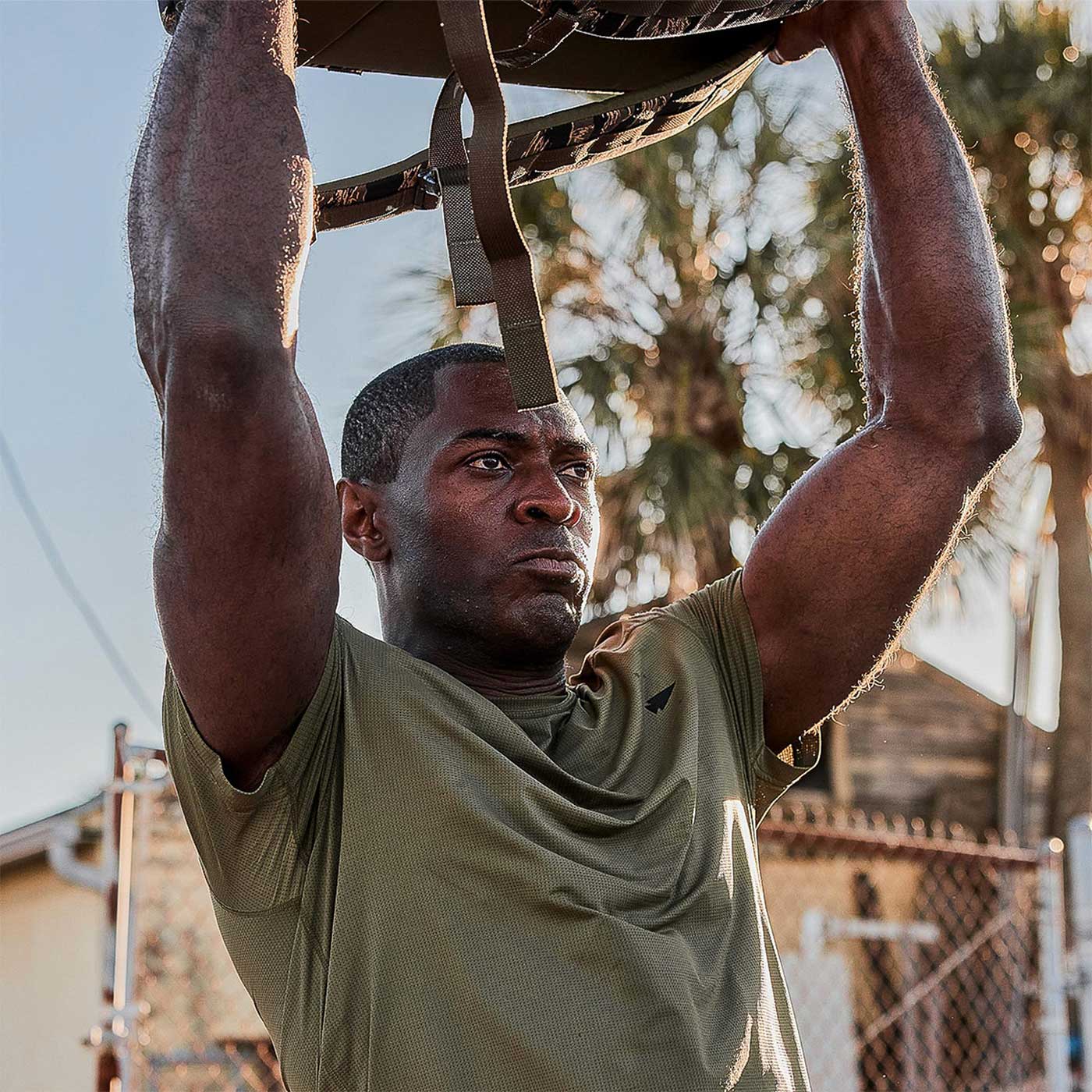 Man in green shirt lifting a sandbag overhead outdoors with palm trees and a fence in the background.