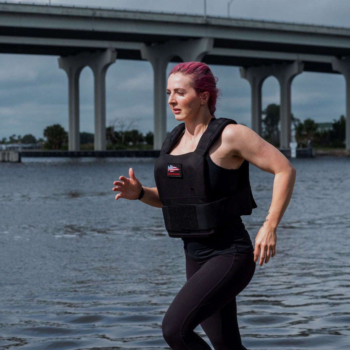 A person with pink hair runs by the water wearing a weighted vest, with a bridge in the background.