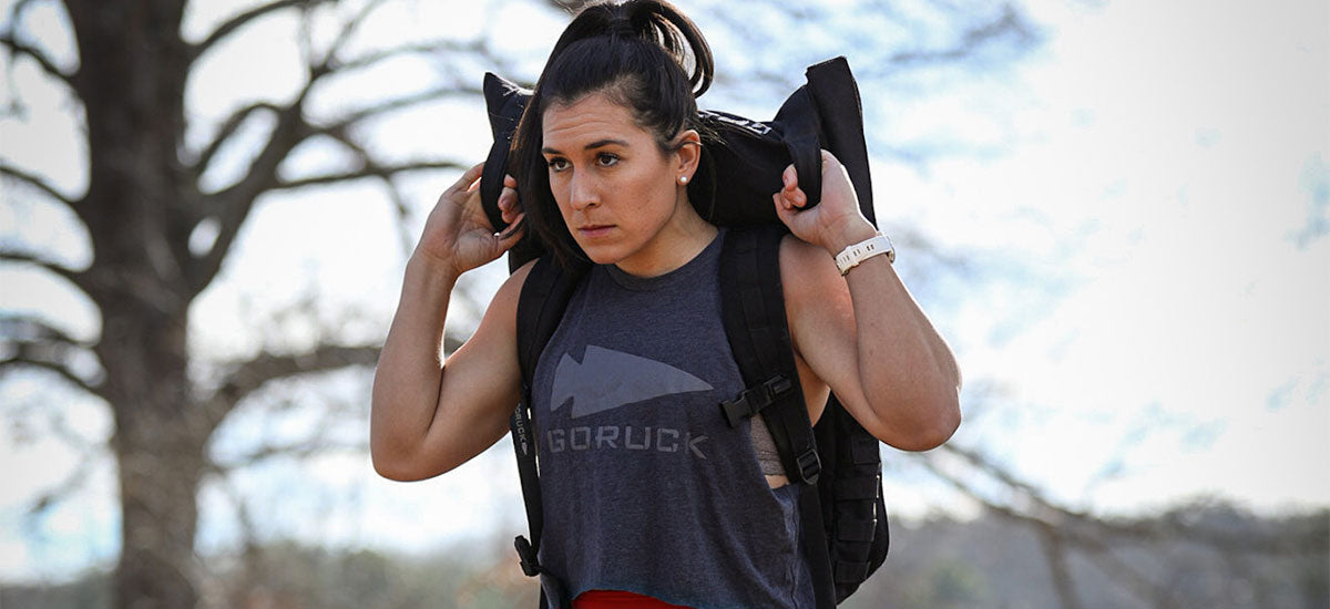 Woman outdoors carrying a weighted backpack on her shoulders, wearing a sleeveless GORUCK shirt.