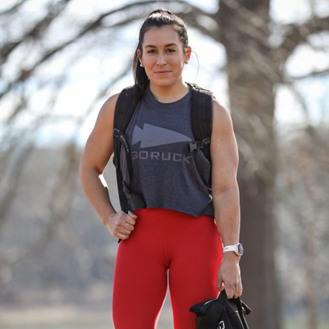 Woman outdoors wearing a sleeveless gray top, red leggings, and a backpack, standing and smiling.