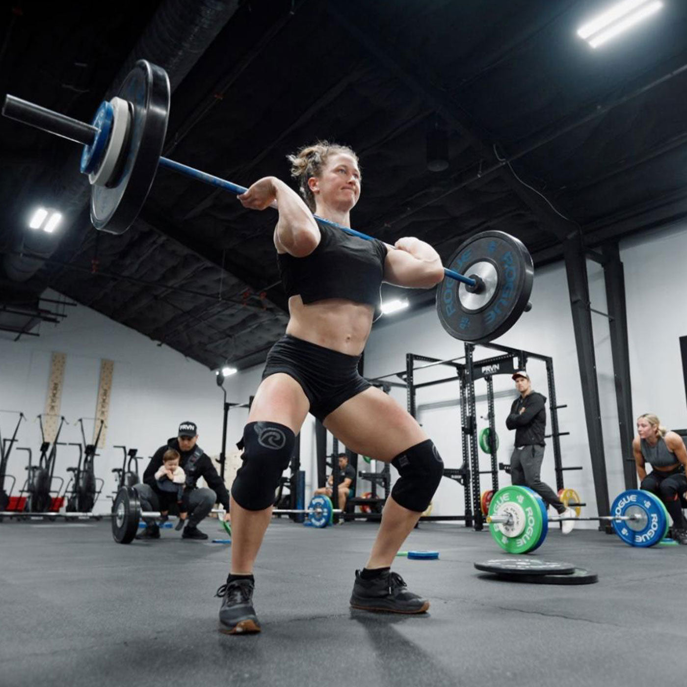 Woman in gym clothes lifting a barbell overhead in a gym with people and equipment in the background.