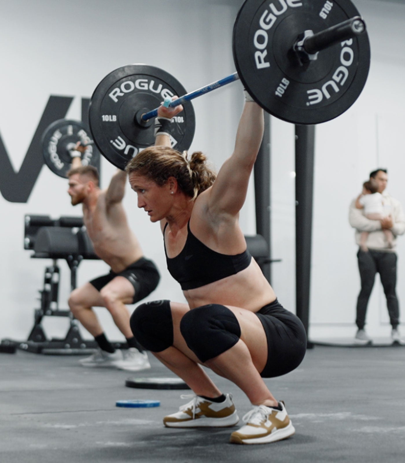 Woman in gym squats with a barbell overhead; another man lifts behind her, people stand in the background.