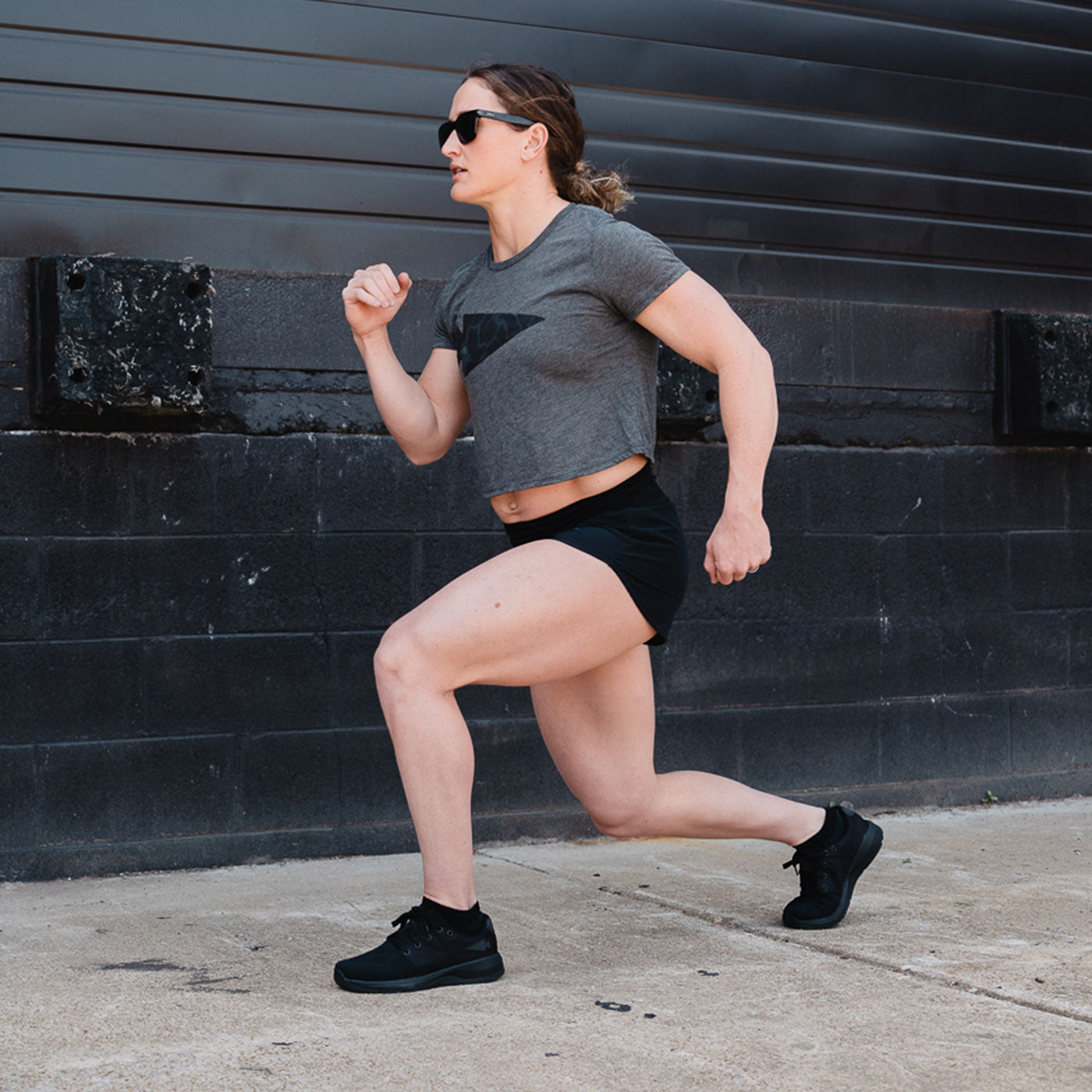 Woman in athletic wear doing a lunge exercise outdoors on a concrete surface near a black wall.