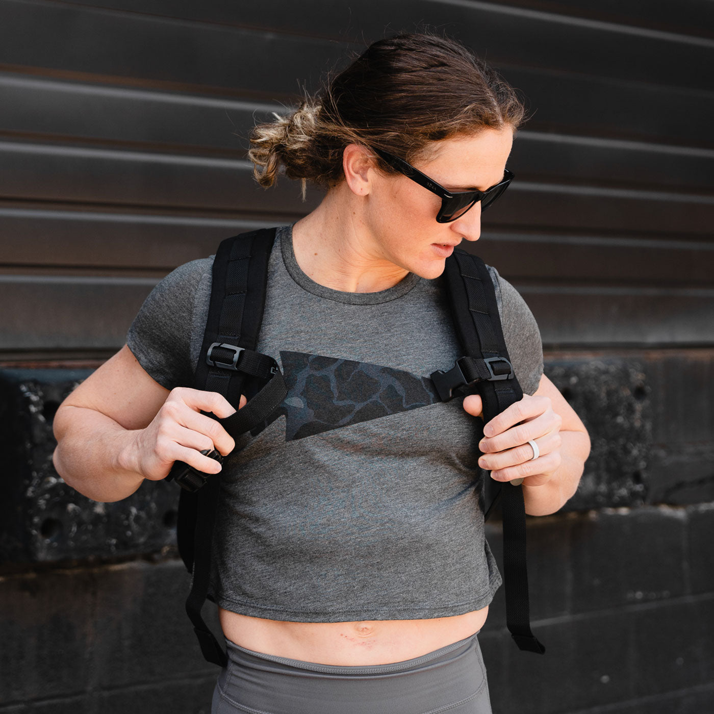 Person in sunglasses adjusts black backpack straps, wearing a gray crop top, standing by a dark metal wall.