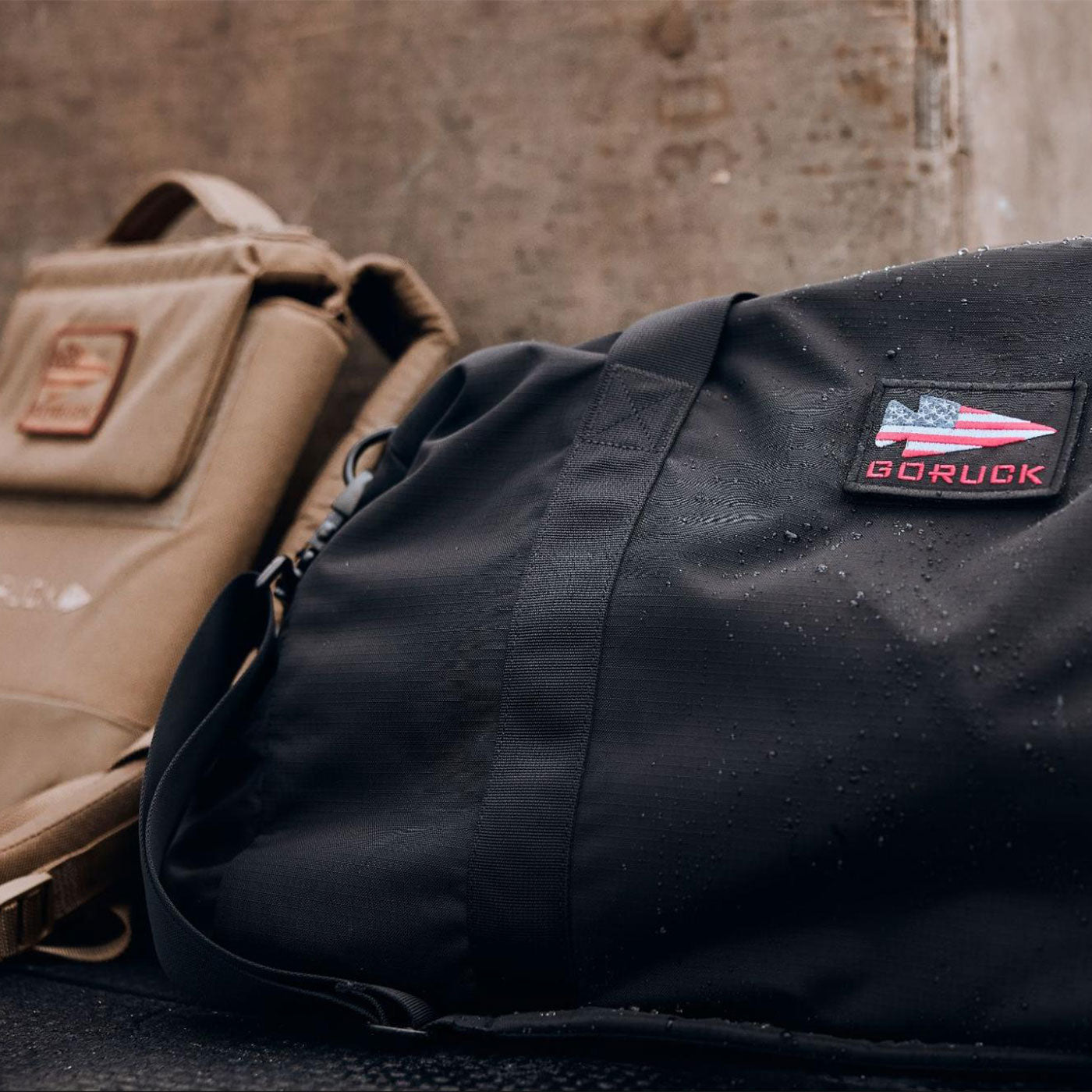 A black duffel bag with a GORUCK patch beside a tan backpack, both on a wet surface.