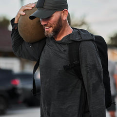 Man wearing GORUCK ToughMesh charcoal hoodie, cap, and backpack carrying a sandbag outdoors