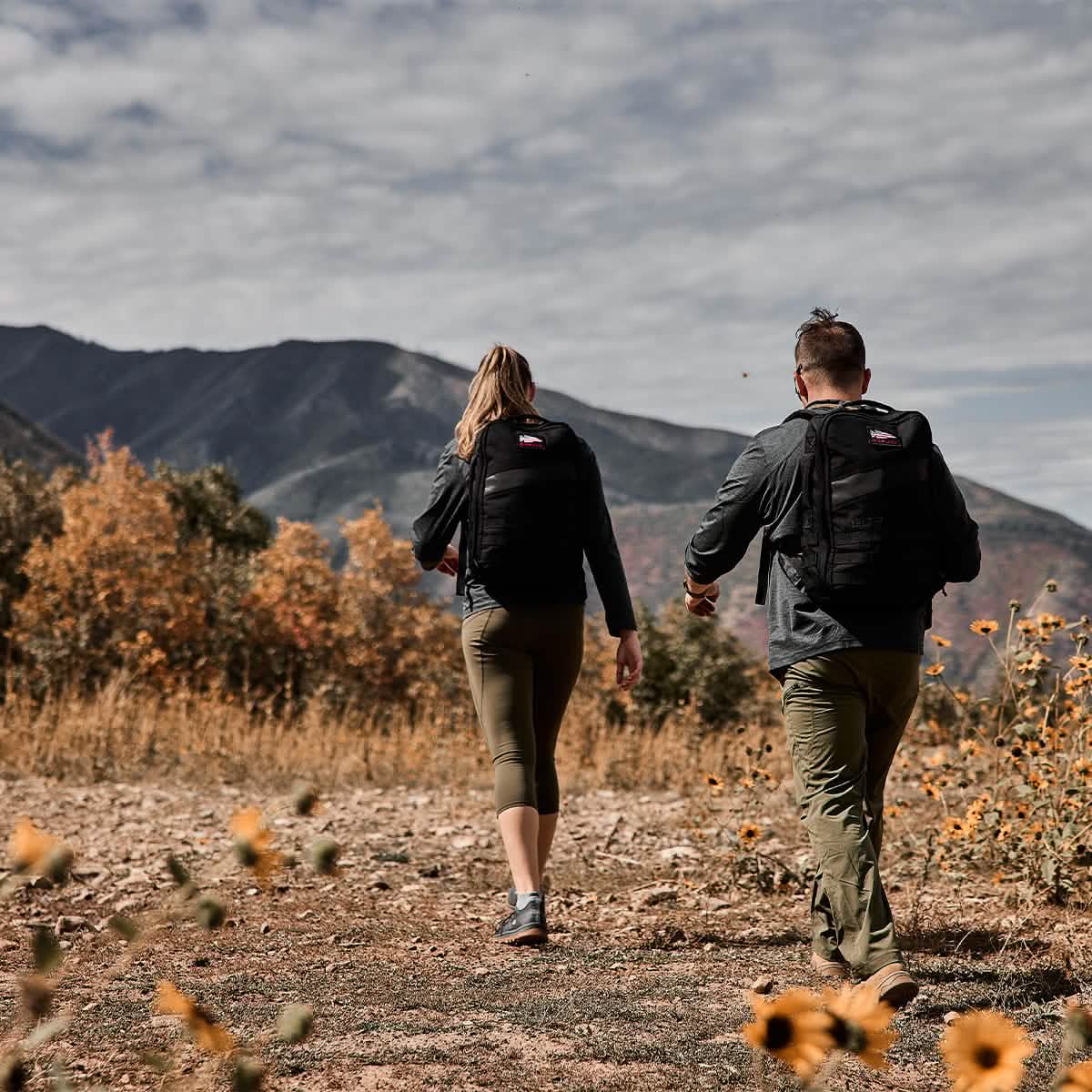 Two people with backpacks hike on a rocky trail with mountains and wildflowers in the background.
