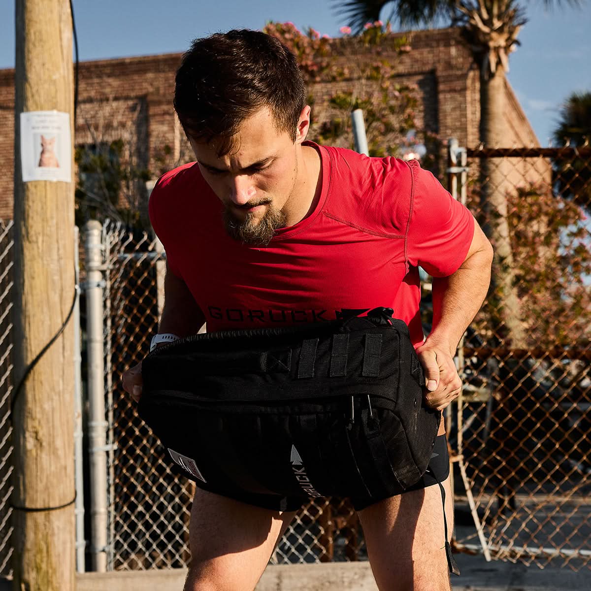 Man in red GORUCK shirt lifting heavy black rucksack outdoors during rucking training
