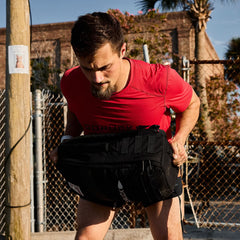 Man in red GORUCK shirt lifting heavy black rucksack outdoors during rucking training