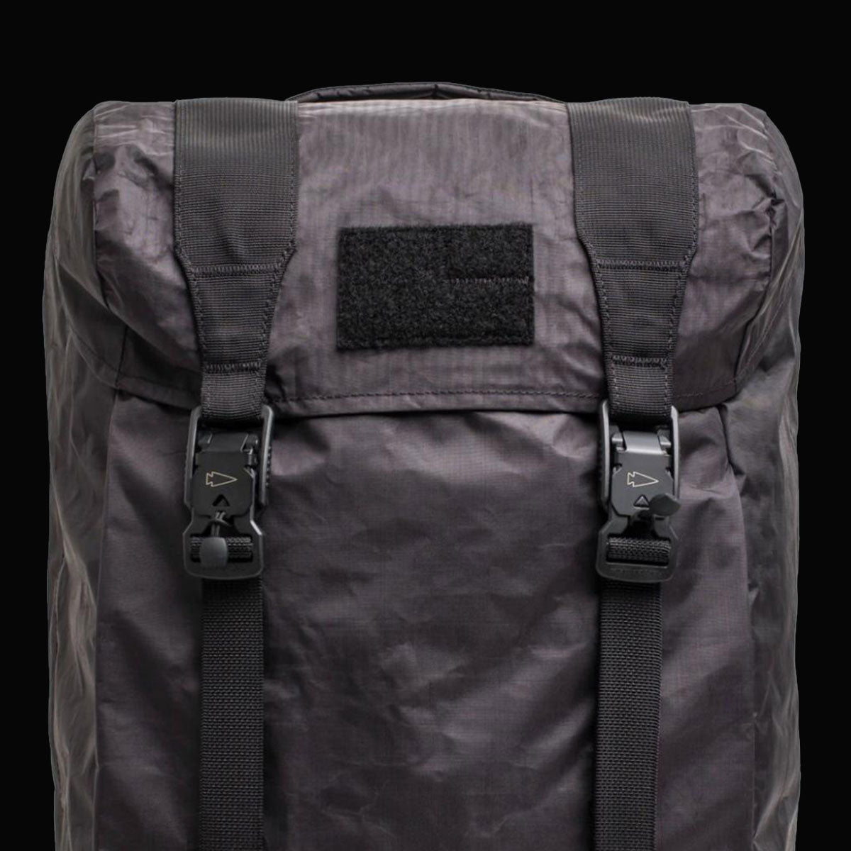 Black backpack with two buckled straps and a rectangular Velcro patch on the front, shown against a black background.