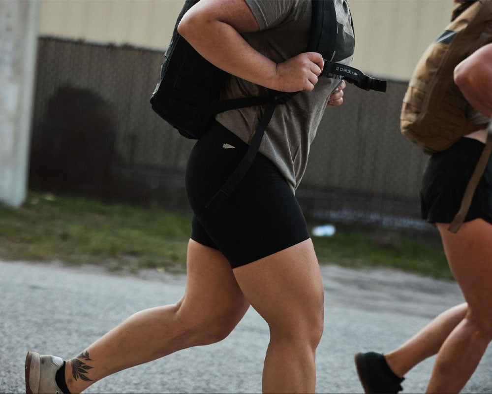 Two people running outdoors wearing backpacks, athletic shorts, and t-shirts, shown from the waist down.