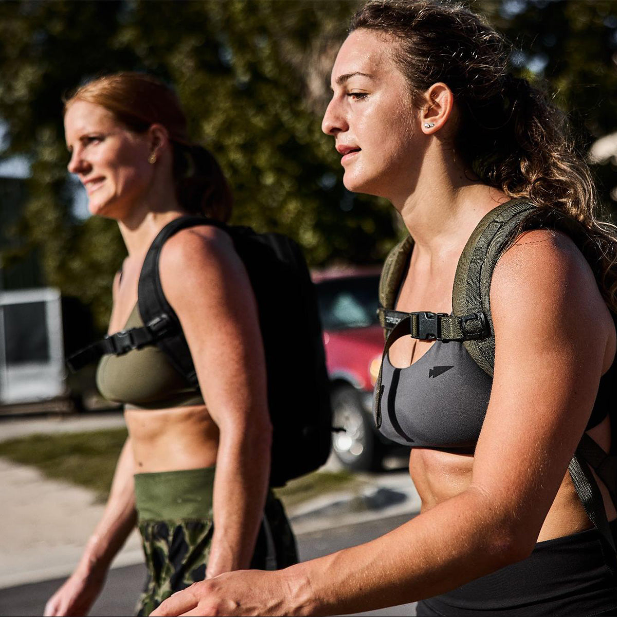 Two women in sports bras and backpacks walk outdoors on a sunny day, appearing focused and active.