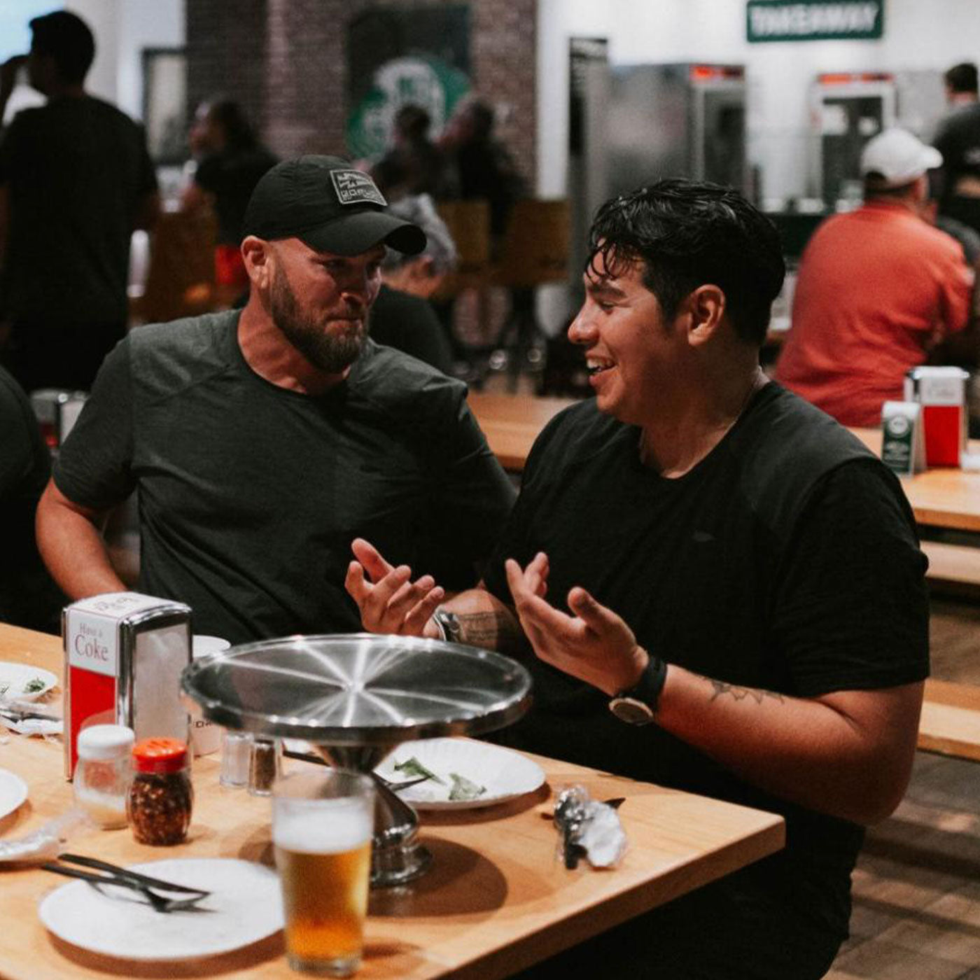 Two men sit at a restaurant table, talking and laughing, with drinks and food in front of them.