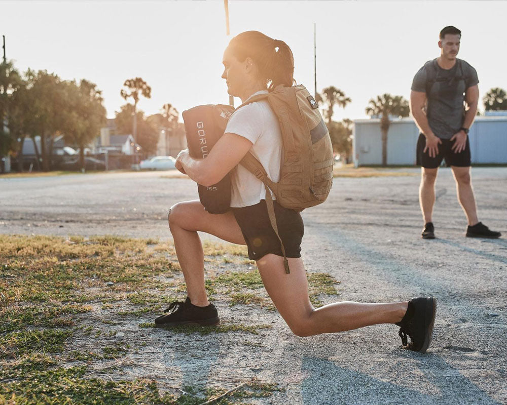Woman lunges outdoors holding a sandbag, wearing a backpack; man stands behind her on a sunny day.