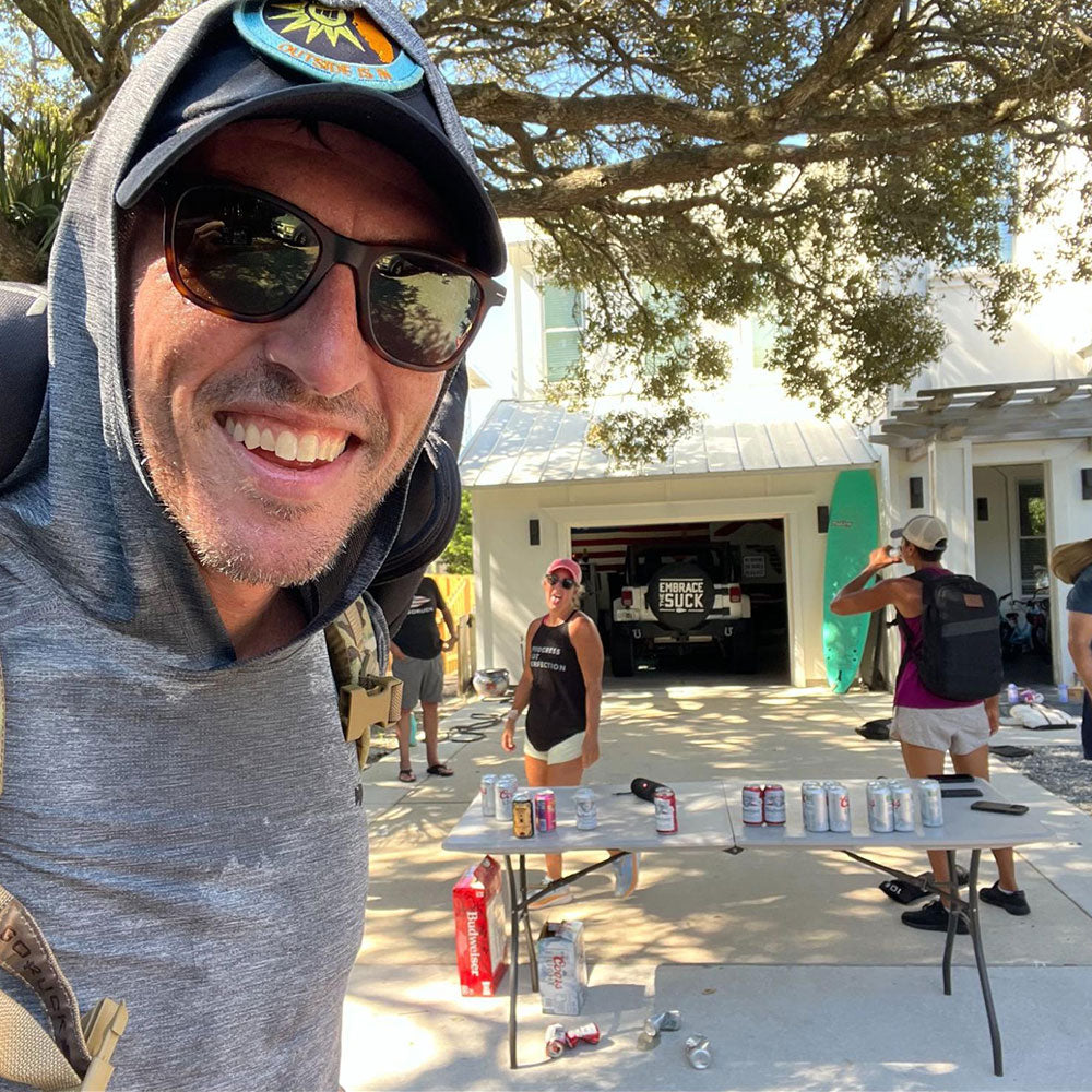 A smiling man in sunglasses takes a selfie outside a house with people and drinks on a table in the driveway.