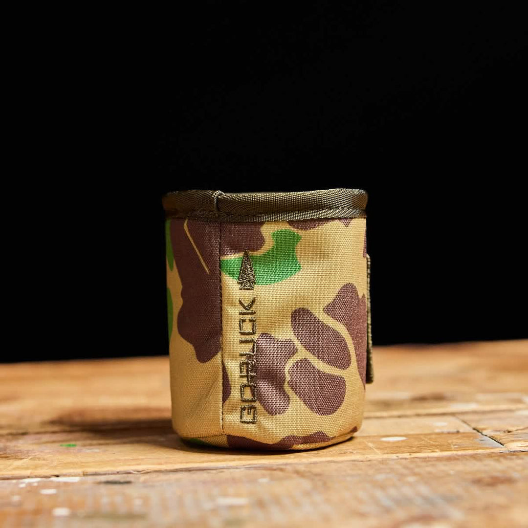 The GORUCK Ballistic Beer Jacket, a compact camo-patterned pouch made from rugged 1000D CORDURA, rests on a wooden surface with a black background.