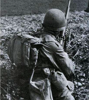 Soldier in combat gear crouches with a rifle, facing away in a grassy, outdoor setting.