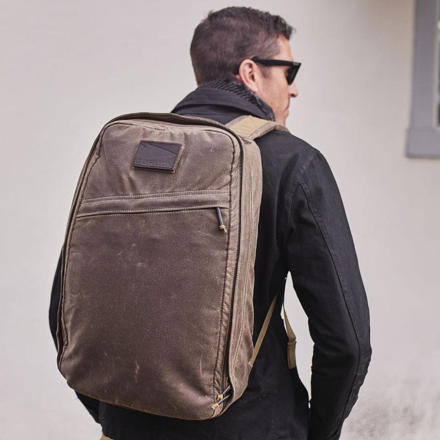 Man wearing sunglasses and a black jacket, carrying a large brown backpack, facing away from the camera.
