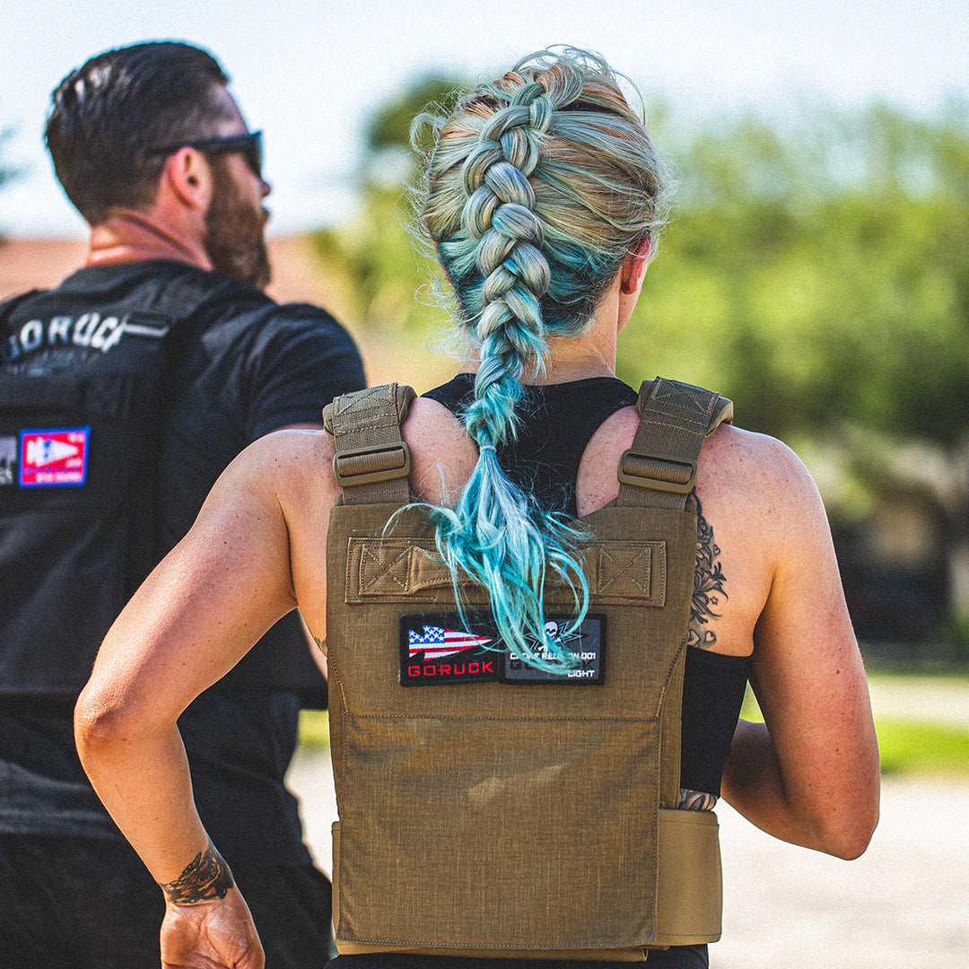 Two people run outdoors wearing GORUCK Training Weight Vest 2.0; one has a long blue braid and visible arm tattoos.