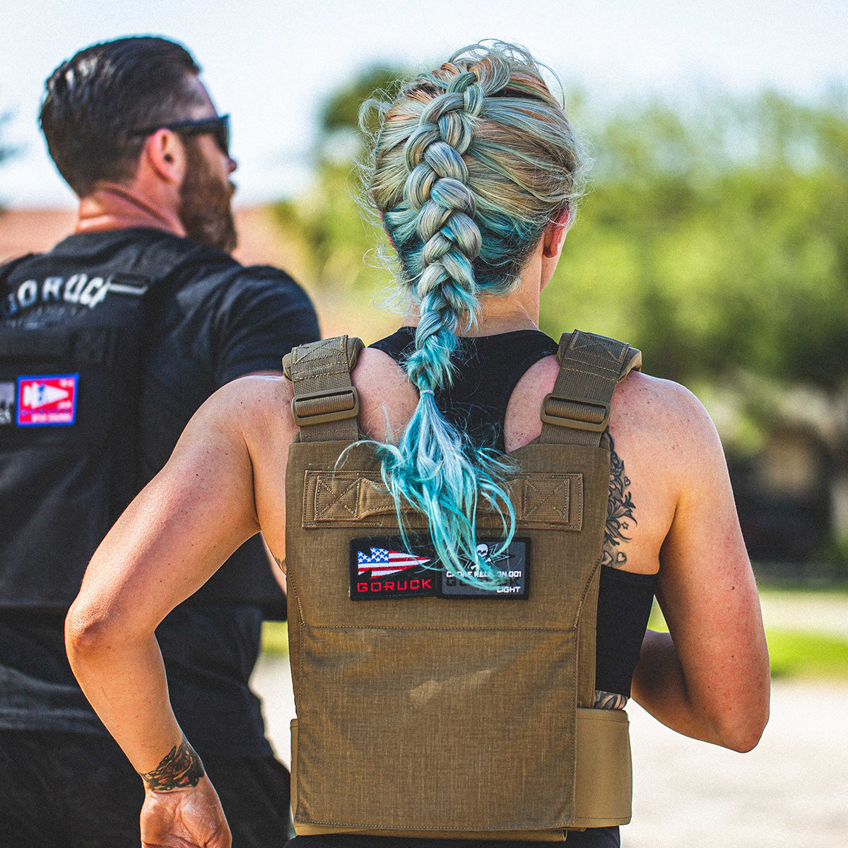 Two people run outdoors wearing GORUCK Training Weight Vest 2.0; one has a long blue braid and visible arm tattoos.
