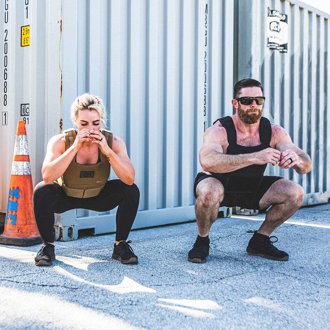 Two people in GORUCK Training Weight Vest 2.0 perform squats outdoors beside a large shipping container.
