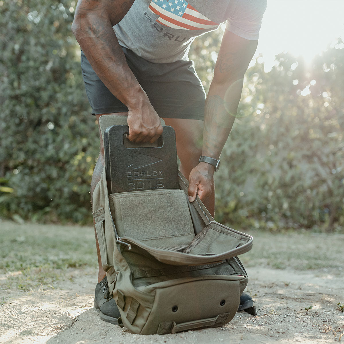 A person places a 30 lb weighted plate into an open backpack outdoors on a sunny day.