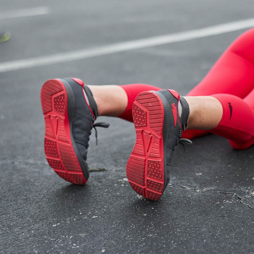 A person in red workout clothes sits on the pavement, highlighting GORUCK Ballistic Trainers in Wolf Grey + High Risk Red W / Red Reflective Spearhead—built with CORDURA® Ballistic Nylon for 3X Stability and functional fitness.