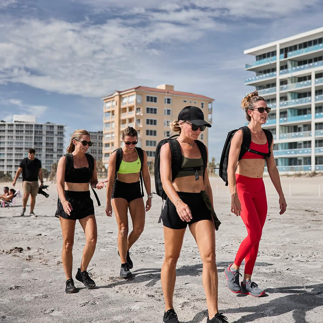 Four women wearing GORUCK Ballistic Trainers in Wolf Grey + High Risk Red stroll along a sandy beach near tall buildings on a sunny day.