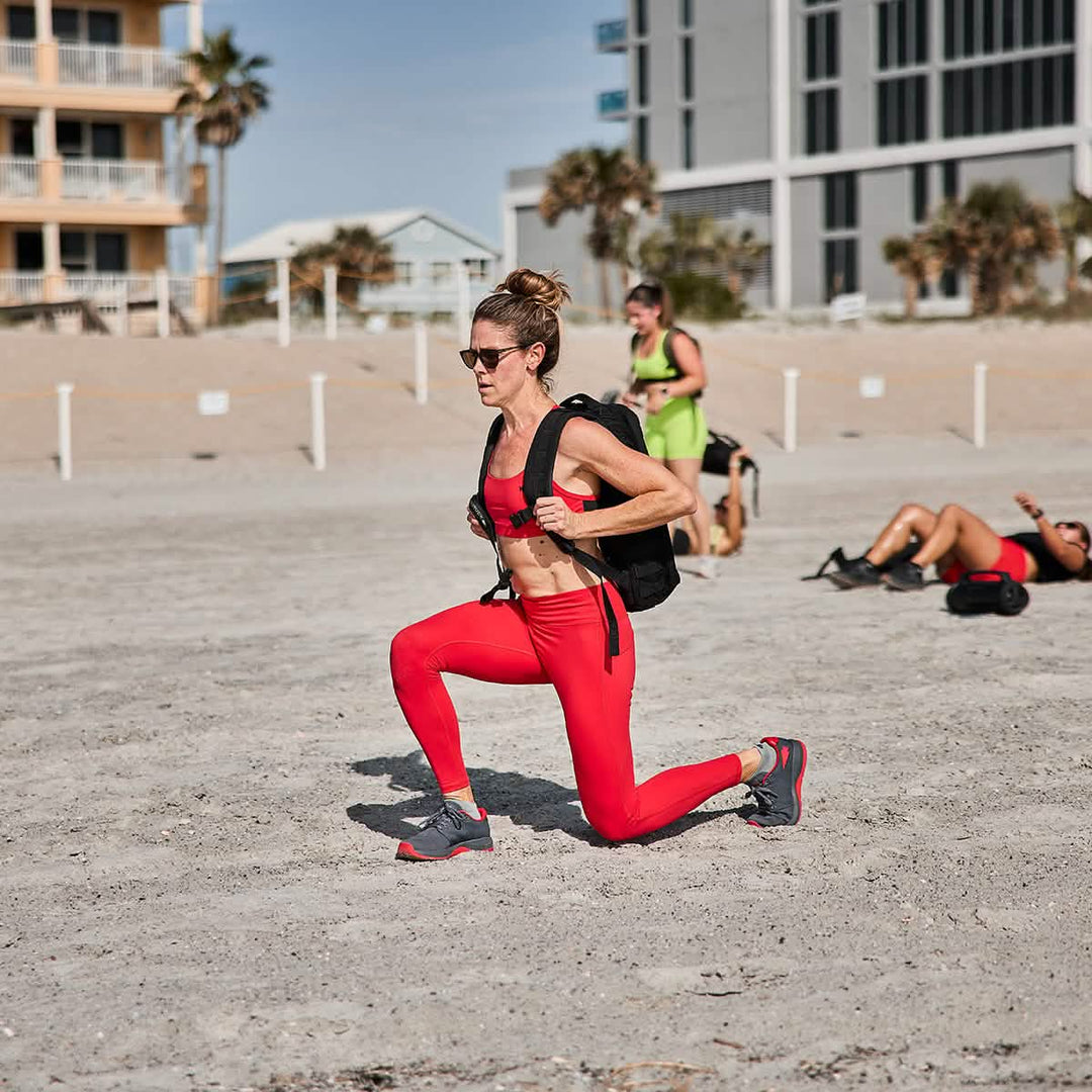 A woman in red workout gear lunges on a sandy beach with buildings behind her, showcasing GORUCK Ballistic Trainers - Wolf Grey + High Risk Red W / Red Reflective Spearhead.