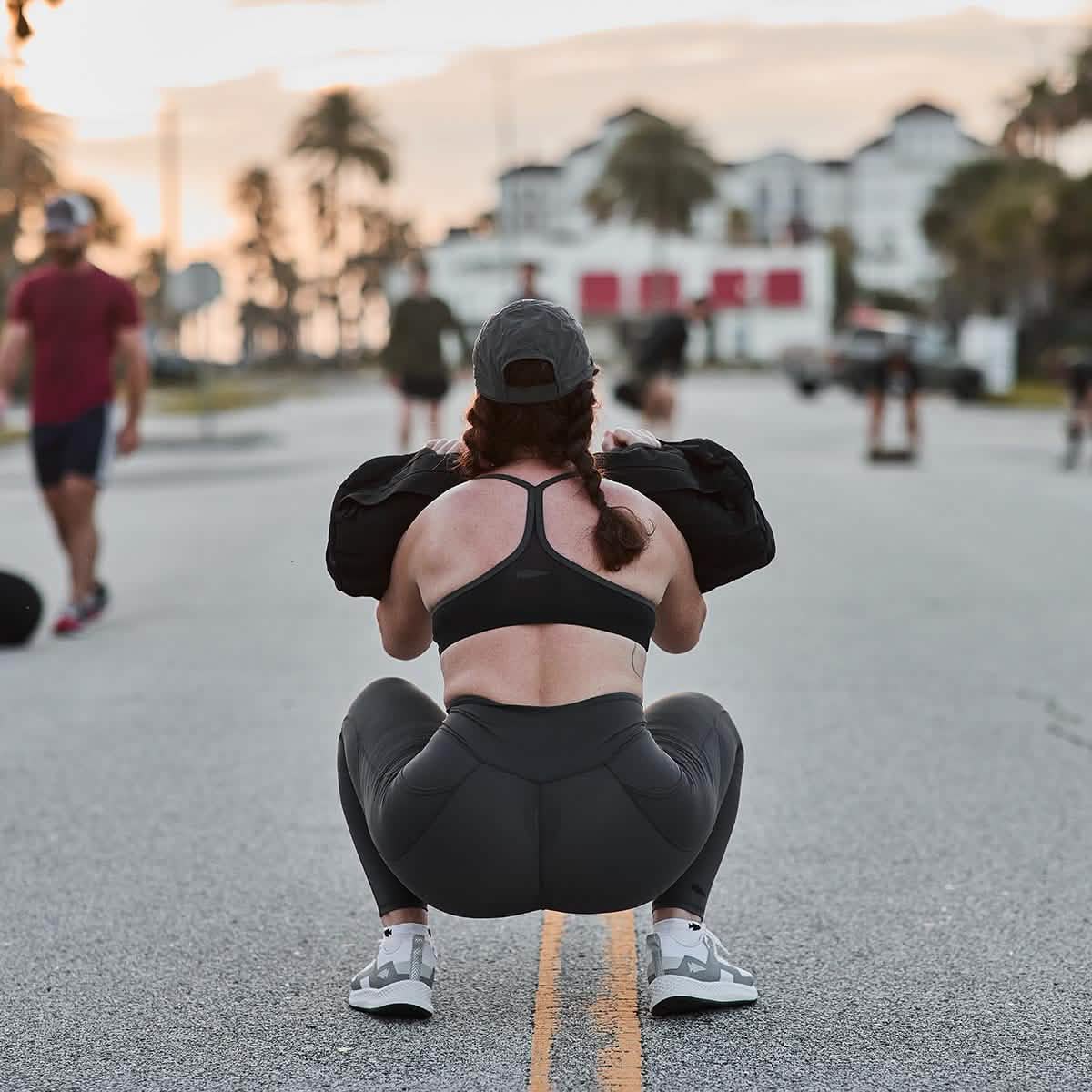 A woman squats on a street, lifting a sandbag in her GORUCK Women’s Training Leggings Pocket USA, with people and palm trees in the background.