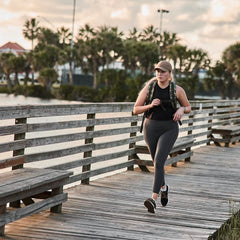 A woman in athletic wear walks on a wooden boardwalk by the water with palm trees, wearing GORUCK Women’s Training Leggings Pocket USA for a comfortable, flexible fit.