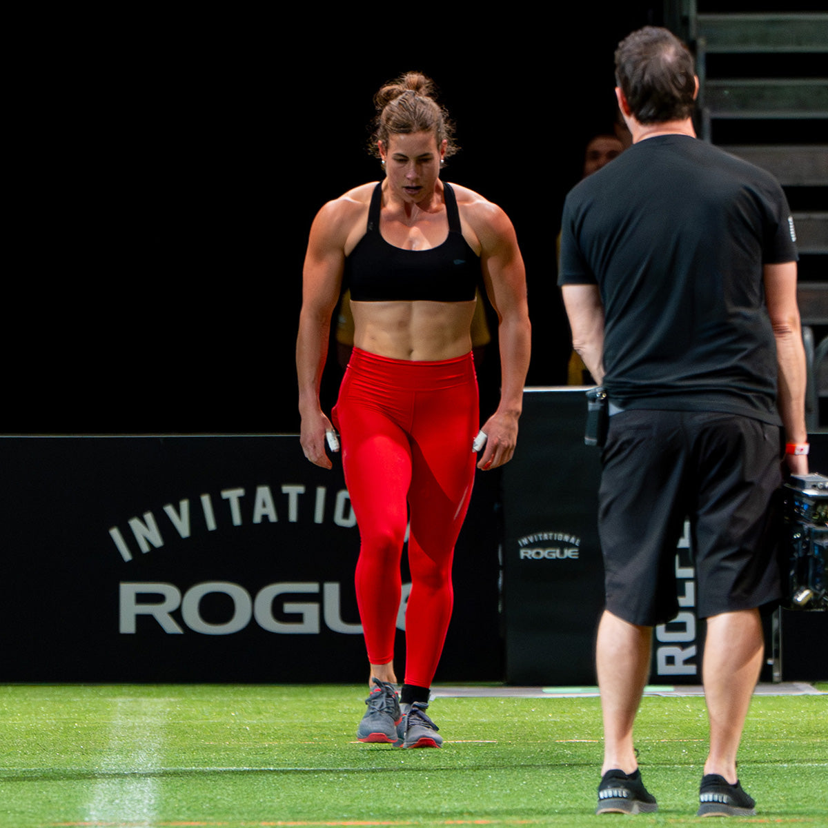 A female athlete in red leggings prepares to compete as a man stands nearby on a sports field.