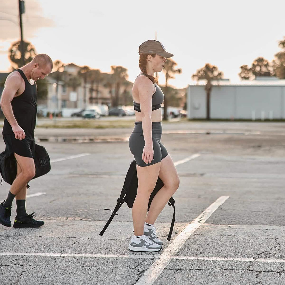 A woman in GORUCK's ToughFlex Women’s Biker Shorts and a cap strolls through an empty parking lot with a man at sunrise. She carries a backpack, highlighting the convenience of stay-put technology, while he looks down, holding his own bag. Trees and buildings create a serene backdrop.