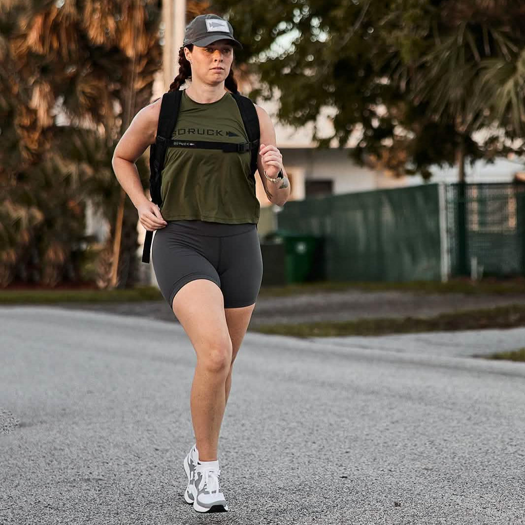 A person in athletic gear is jogging on a paved road, surrounded by trees and greenery. They are wearing a cap and Women’s Biker Shorts made of ToughFlex fabric from GORUCK, focusing on the path ahead. It’s a sunny day, creating a warm ambiance.