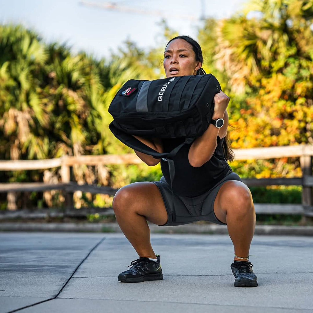 Surrounded by lush greenery and a wooden fence, a person squats with a heavy backpack, highlighting the indestructible ToughFlex Women’s Biker Shorts by GORUCK. Clad in athletic wear and wearing a watch, they depend on the shorts' stay-put technology to maintain perfect form during their workout.