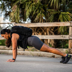 A woman in athletic wear performs a plank exercise on a paved surface, demonstrating the stay-put technology of her GORUCK Women’s Biker Shorts - ToughFlex. She is wearing a backpack and sneakers. In the background, there is a wooden fence and palm trees.